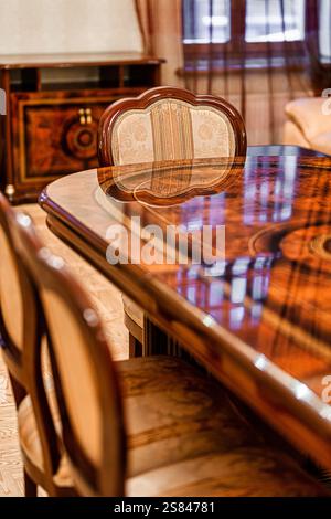 Una raffinata sala da pranzo presenta un tavolo in legno lucido, sedie imbottite con disegni ornati, un armadietto dettagliato e una finestra con tende trasparenti. Foto Stock