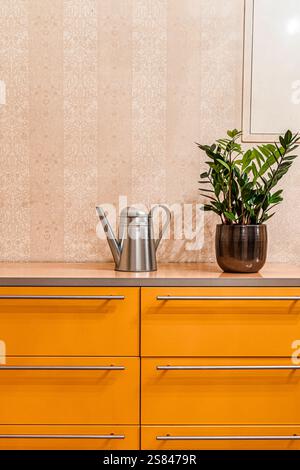 Un armadietto giallo con maniglie argentate contiene una tanica metallica e una pianta in vaso verde. La carta da parati floreale beige aggiunge texture allo sfondo. Foto Stock