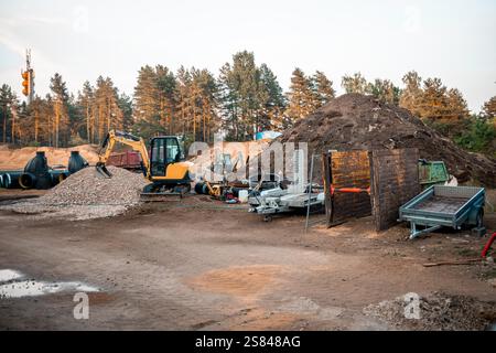 Un cantiere in attività con un escavatore giallo, un mucchio di ghiaia, un tumulo di terra, tubi a spirale e macchinari, delimitata da alberi alti sotto una s limpida Foto Stock