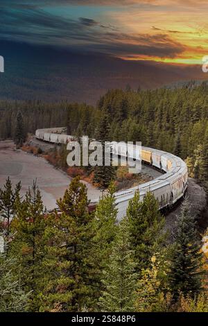 Un treno merci si snoda attraverso una lussureggiante foresta al tramonto, circondato da vibranti colori autunnali e da una natura selvaggia panoramica Foto Stock