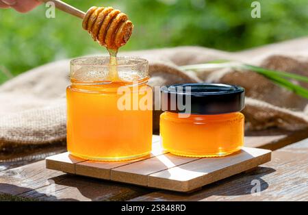 Il miele dorato fresco viene gocciolato da un cucchiaio in un vaso di vetro, collocato all'aperto in una giornata di sole, evidenziandone la dolcezza naturale. Foto Stock