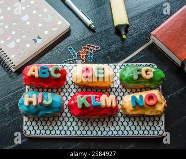 Sei eclari colorati con glassa alfabetica si trovano su un piatto a motivi geometrici, circondati da un notebook, una penna, un evidenziatore, graffette e un diario in pelle. Foto Stock