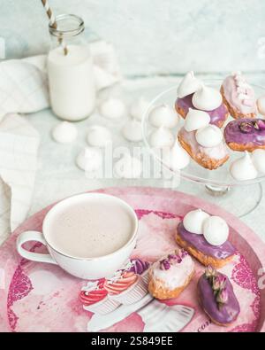 Un dessert a tema pastello con eclari viola ghiacciati, meringhe bianche, un caffè schiumoso su un vassoio rosa e una bottiglia di latte con cannuccia a righe. Foto Stock