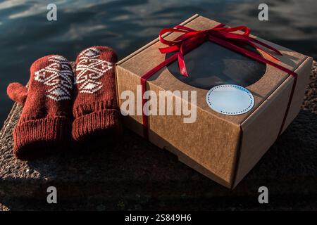 Un paio di guanti rossi con motivi bianchi giace accanto a una confezione regalo marrone con un nastro rosso su una superficie di pietra vicino a ondulazioni di acqua scura. Foto Stock