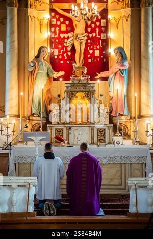 Un altare religioso all'interno di una chiesa con un crocifisso, statua della Vergine Maria, accenti d'oro, candele e membri del clero inginocchiati nella preghiera. Foto Stock
