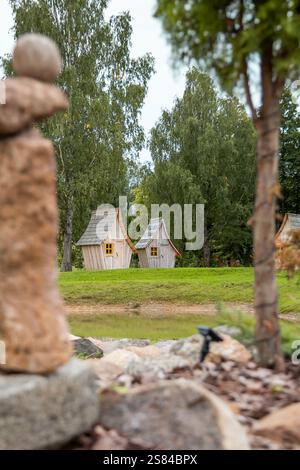 Due case in legno con tetti curvi e finestre con cornici gialle si trovano vicino a uno stagno, circondate da alti alberi verdi, erba e rocce in un ambiente naturale Foto Stock