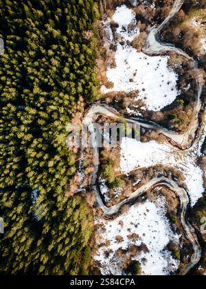 Una tortuosa strada di montagna attraversa una fitta foresta sempreverde da un lato e un terreno innevato dall'altro, mostrando contrasti stagionali. Foto Stock