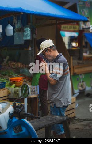 Balikpapan, Indonesia - 25 dicembre 2024. Il vecchio con un berretto e un bicchiere sta accendendo una sigaretta al mercato. Foto Stock