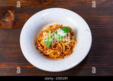 Un piatto di spaghetti condito con una ricca salsa di carne e basilico fresco è posto su un rustico tavolo di legno. Il piatto è guarnito con formaggio grattugiato, invit Foto Stock