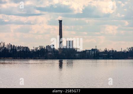 L'immagine raffigura un tranquillo paesaggio lungo il fiume con una struttura industriale e un alto camino sullo sfondo, circondato da alberi senza foglie sotto un par Foto Stock