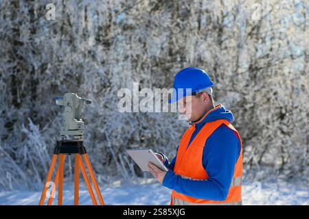 Surveyor che utilizza tablet e teodolite durante l'inverno topografia in un paesaggio innevato. Foto Stock