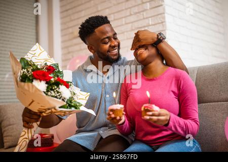 Felice coppia amorevole che festeggia il compleanno della donna a casa. Il fidanzato sta dando un bouquet di fiori come sorpresa alla sua ragazza. Foto Stock