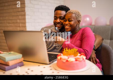 Felice giovane coppia che festeggia il compleanno della donna a casa. Stanno brinando con del vino e stanno facendo videochiamate con la famiglia. Foto Stock