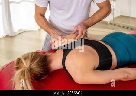 Osteopata maschile irriconoscibile che guarda in basso mentre si è in piedi vicino a un tavolo con paziente donna anonima sdraiata e che esegue un massaggio miofasciale della scapola Foto Stock