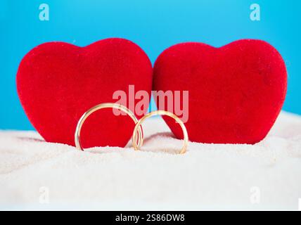 Due cuori di velluto rosso su uno sfondo di sabbia con un cielo blu, accompagnati da fedi nuziali dorate. Perfetto per i temi di San Valentino, i matrimoni Foto Stock