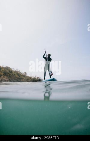 Un uomo è in piedi su una tavola da surf nell'oceano. Sta tenendo una pagaia e si trova nel mezzo di un'onda. La scena è calma e pacifica, come l'uomo è enj Foto Stock