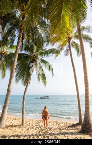 Giovane donna bionda in piedi sotto le palme e godersi il sole mattutino sulla spiaggia tropicale di Maenam a Koh Samui, Thailandia. Foto Stock