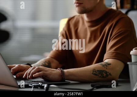 Foto ritagliata di uno sviluppatore di software maschile irriconoscibile con tatuaggi che scrivono sul laptop mentre lavora nell'ufficio dell'azienda IT, spazio di copia Foto Stock