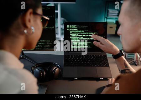 Immagine ritagliata di un programmatore IT maschile irriconoscibile che punta sullo schermo del notebook con righe di codice in esecuzione collaborando con una collega donna che lavora fino a tardi in ufficio Foto Stock
