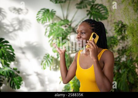 Una donna nera interessata si prende una pausa per chattare con lo smartphone hanno una piacevole conversazione circondata da piante. Foto Stock
