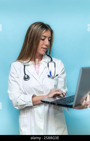 Il medico donna verticale che indossa un cappotto bianco e uno stetoscopio sta lavorando con il computer portatile su sfondo blu, digitando sulla tastiera e guardando lo schermo, concetto Foto Stock