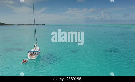 I turisti che si godono un'avventura in barca a vela nella laguna turchese di Bora Bora, scivolando oltre i bungalow sull'acqua sotto un cielo luminoso, catturando l'essenza del paradiso tropicale nella Polinesia francese Foto Stock