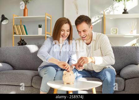 Felice giovane coppia seduta sul divano, risparmiando soldi nel salvadanaio, discutendo piani finanziari a casa. Foto Stock