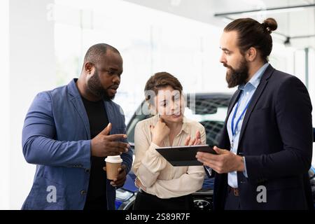 Il venditore della concessionaria tiene in mano un tablet per rispondere alle domande dei clienti sulle auto, sul finanziamento e sul processo di acquisto. L'agente dello showroom parla con i clienti delle garanzie e dei servizi che utilizzano il dispositivo Foto Stock