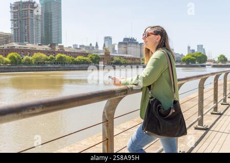 Giovane donna sorridente mentre si appoggia su una ringhiera vicino al fiume, skyline urbano alle sue spalle in una giornata di sole. Foto Stock