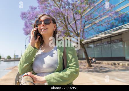 Donna d'affari che usa lo smartphone vicino alla fioritura dell'albero jacaranda mimosifolia, godendo di una giornata di sole in ambiente urbano Foto Stock