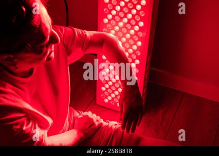 Donna anziana seduta sul pavimento tenendo il braccio vicino a un dispositivo terapeutico a luce rossa Foto Stock