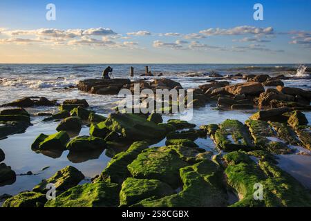 Una vista panoramica di una costa rocciosa con vivaci pietre verdi ricoperte di muschio, onde che si infrangono sullo sfondo e un fotografo che cattura la bellezza Foto Stock