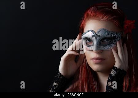 Ritratto di una bella giovane donna con i capelli rossi che ricoprono parte del suo viso con la maschera colombina. Tutto su sfondo nero. Orizzontalmente. Foto Stock