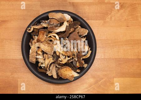 Colpo di testa di funghi selvatici secchi assortiti in una ciotola di ceramica scura su sfondo di legno naturale. Foto Stock