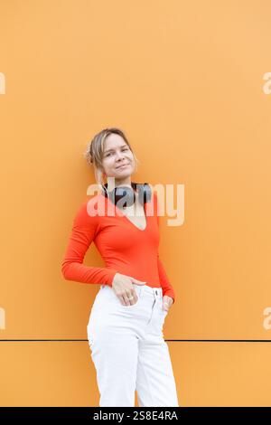 Una donna con il top rosso e i pantaloni bianchi si appoggiano su una parete arancione a Varsavia, con le cuffie intorno al collo lo sfondo vivace migliora il suo stato di salute Foto Stock