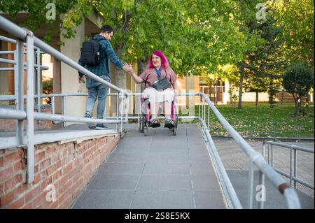 Una coppia al college, con la donna che usa una sedia a rotelle a causa della spina bifida, navigano insieme nel campus, mostrando accessibilità e supporto Foto Stock