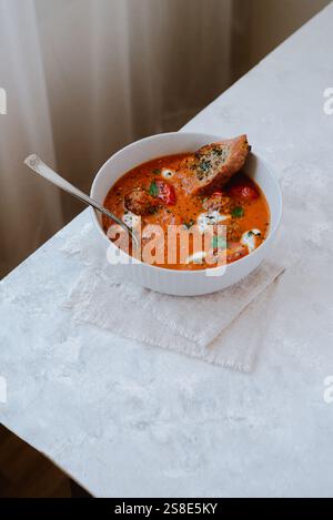 Vista dall'alto di una zuppa di pomodoro saporita in una ciotola bianca, con basilico, panna, polpette, pomodori ciliegini, e una fetta di pane tostato. Foto Stock