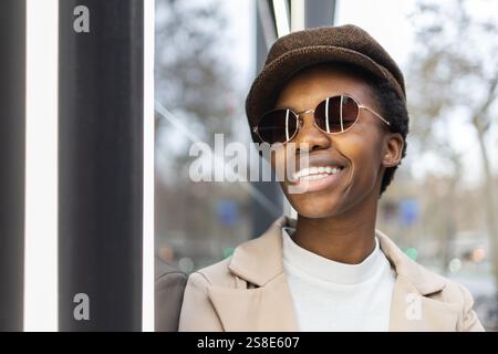Donna nera alla moda che indossa occhiali da sole e un cappello, sorridendo con sicurezza in un ambiente urbano, il suo abbigliamento elegante e i suoi accessori esaltano la sua vibra Foto Stock
