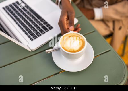 Una donna di colore tiene un cappuccino mentre lavora su un notebook in un bar all'aperto, la scena cattura un mix di lavoro e svago, riflettendo un'atmosfera vivace Foto Stock