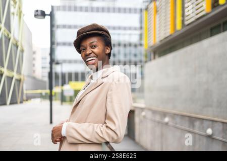 Un'elegante donna nera sorride con fiducia mentre si trova in un ambiente urbano moderno il suo abbigliamento chic e il suo comportamento gioioso riflettono l'empowerment e l'eleg Foto Stock
