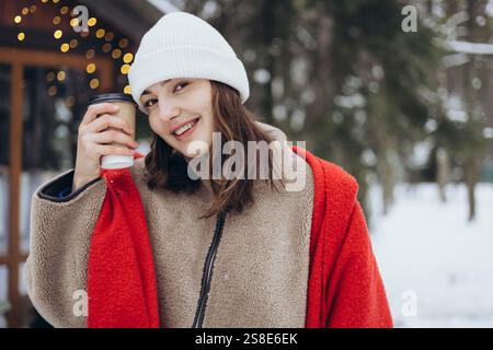 Una giovane donna sorride tenendo in mano una tazza di caffè, vestita calorosamente con una sciarpa rossa e berretto bianco su uno sfondo innevato con luci soffuse. Foto Stock