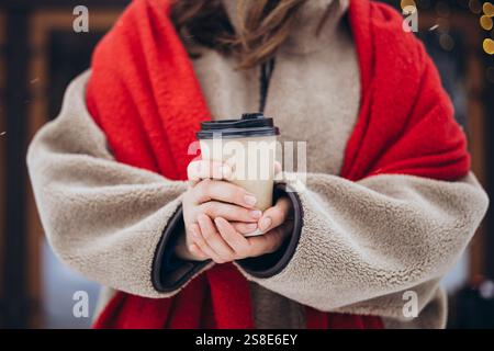 Una giovane donna corta e irriconoscibile, vestita con un comodo cappotto in teddy e una sciarpa rossa che regala una tazza di caffè da asporto, per mantenersi al caldo durante la giornata invernale Foto Stock