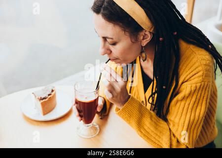 Una donna con dreadlocks in un caffè sorseggia una bevanda attraverso una paglia che indossa un maglione giallo e un cerchietto, si gode una fetta di torta su un legno chiaro Foto Stock