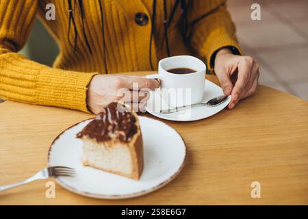 Una persona con un maglione giallo caldo si gode una serena pausa caffè con una tazza di caffè e una deliziosa fetta di torta al cioccolato su un tavolo di legno Foto Stock