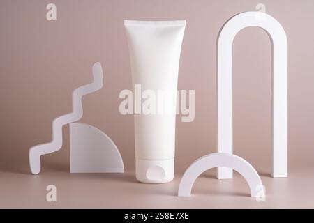 Un concetto minimalista di cura della pelle caratterizzato da un tubo bianco circondato da eleganti forme geometriche su sfondo beige, che incarna semplicità ed eleganza Foto Stock