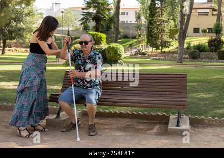 Un uomo anziano ipovedente con occhiali da sole e bastone bianco, seduto su una panchina del parco, riceve l'assistenza della sua figlia di supporto in un floreale Foto Stock