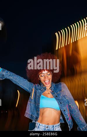 Donna discendente africana con vibranti capelli afro che ballano gioiosamente sotto le luci notturne. Indossando occhiali e una giacca in denim, incarna una vivace città Foto Stock