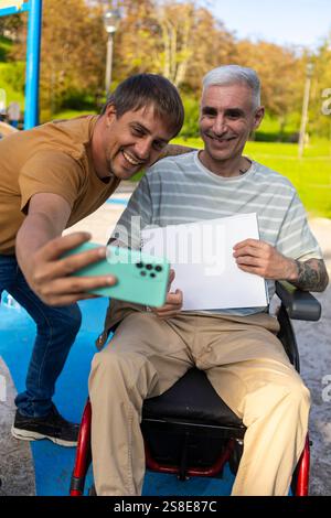 Due amici in un parco si divertono a scattare un selfie insieme una persona è seduta su una sedia a rotelle per paralisi cerebrale, tenendo in mano una pagina vuota del taccuino, mentre il Foto Stock