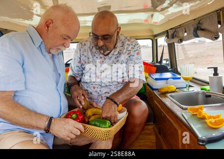 Le coppie gay senior amano preparare un pasto fresco e colorato all'interno del loro veicolo fuoristrada durante un viaggio in campeggio, mostrando un momento di dolce festa Foto Stock
