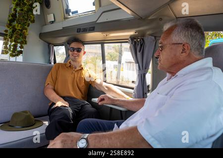 Due uomini, uno più giovane e uno più grande, sono impegnati in una conversazione amichevole all'interno di un camper. L'uomo più giovane sorride e indossa occhiali da sole, whi Foto Stock